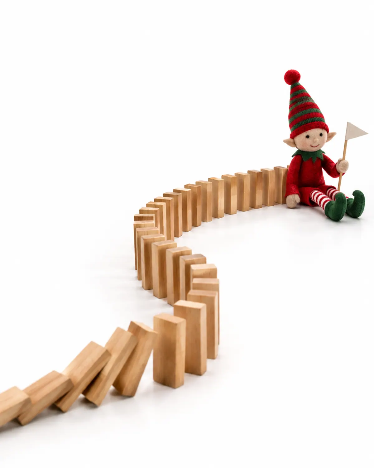 A curving line of wooden dominoes on a living room floor leading to a felt elf doll holding a tiny paper flag