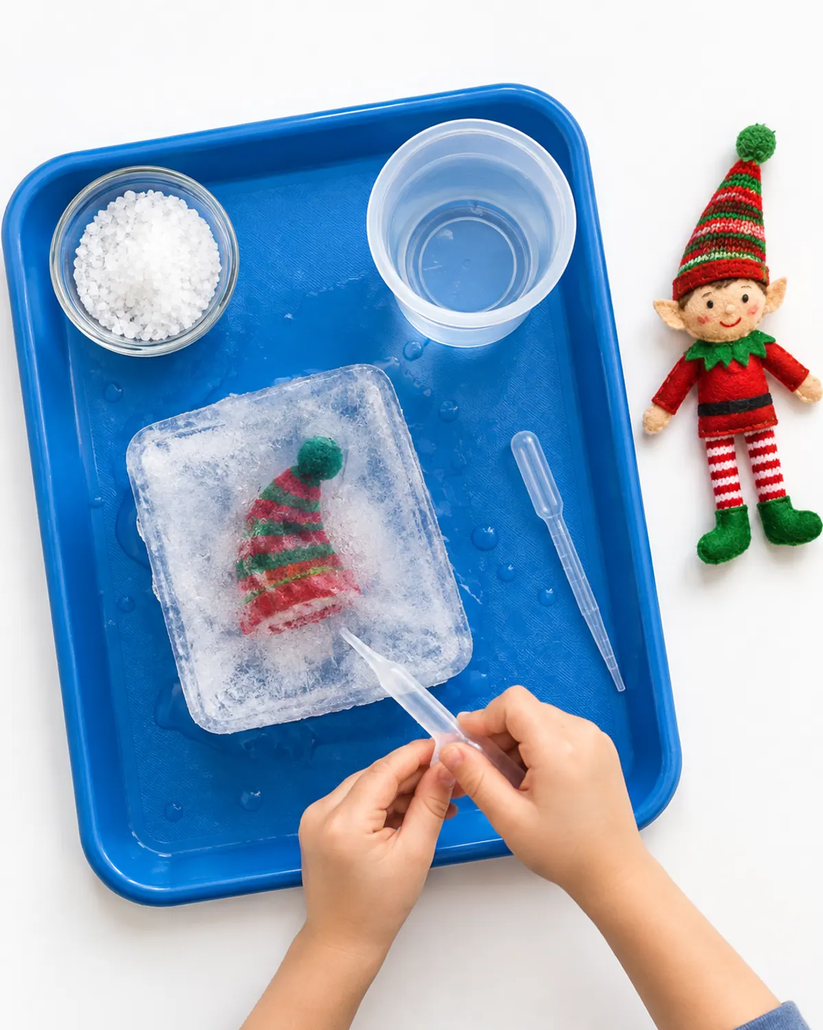 A child using a dropper on a block of ice with a tiny knit elf hat frozen inside, salt and warm water on a tray