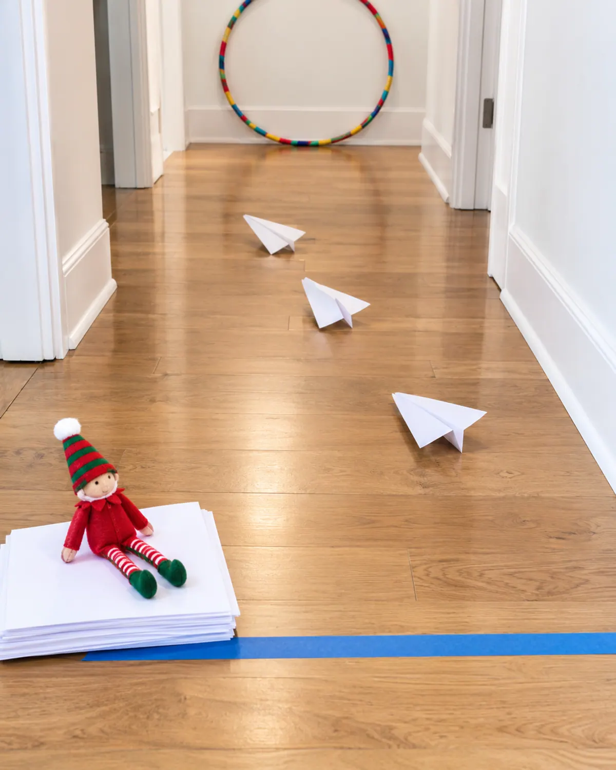 Folded paper airplanes on a hallway floor with a hula hoop target at the far end and an elf doll by the tape start line