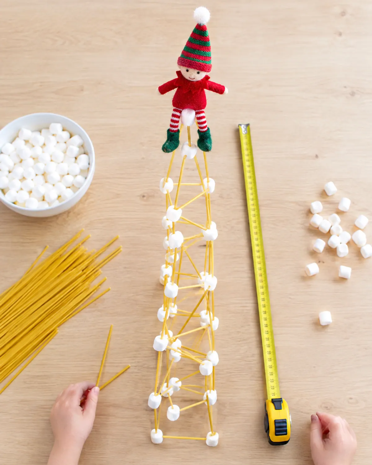 A tall tower of dry spaghetti and mini marshmallows with a felt elf doll balanced on top and a tape measure beside it