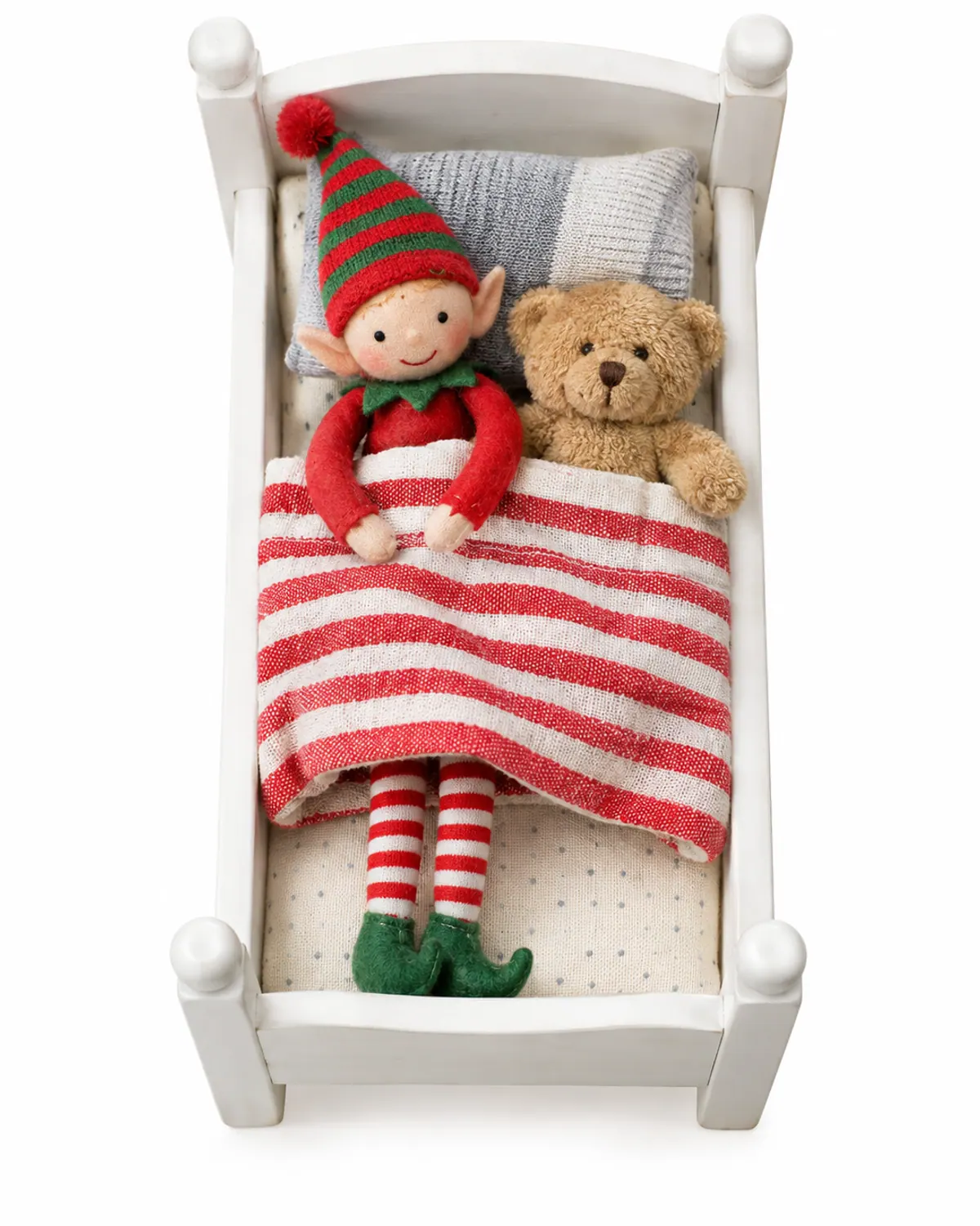 A felt elf doll tucked into a small white doll bed beside a teddy bear under a red-and-white striped blanket, a calm elf idea for toddlers