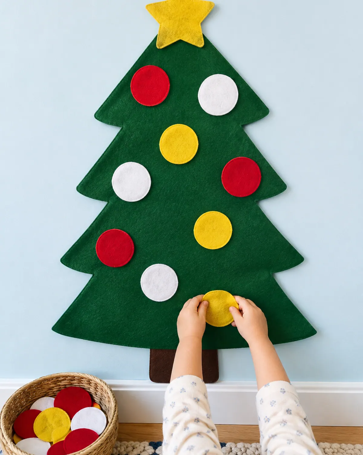 Toddler hand pressing a yellow felt ornament onto a large felt Christmas tree on a nursery wall