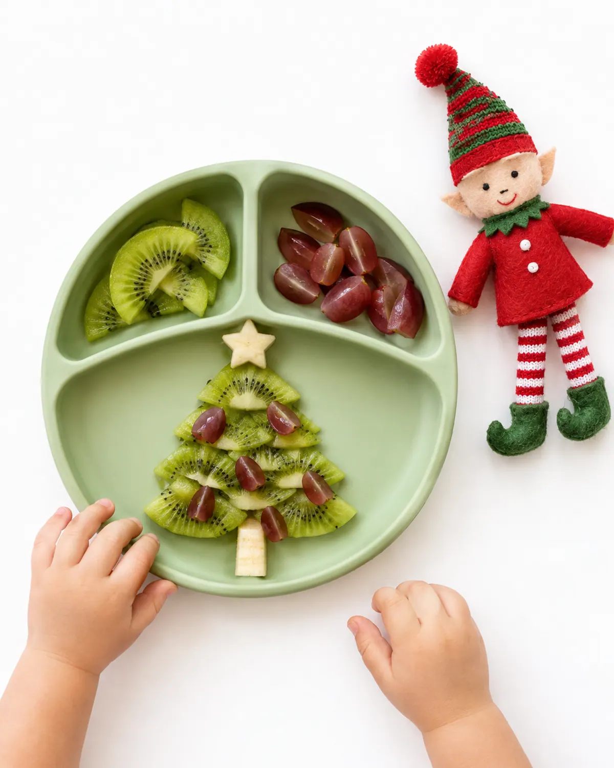 Toddler plate with kiwi slices arranged as a Christmas tree with quartered grapes and a banana trunk, a felt elf doll beside it