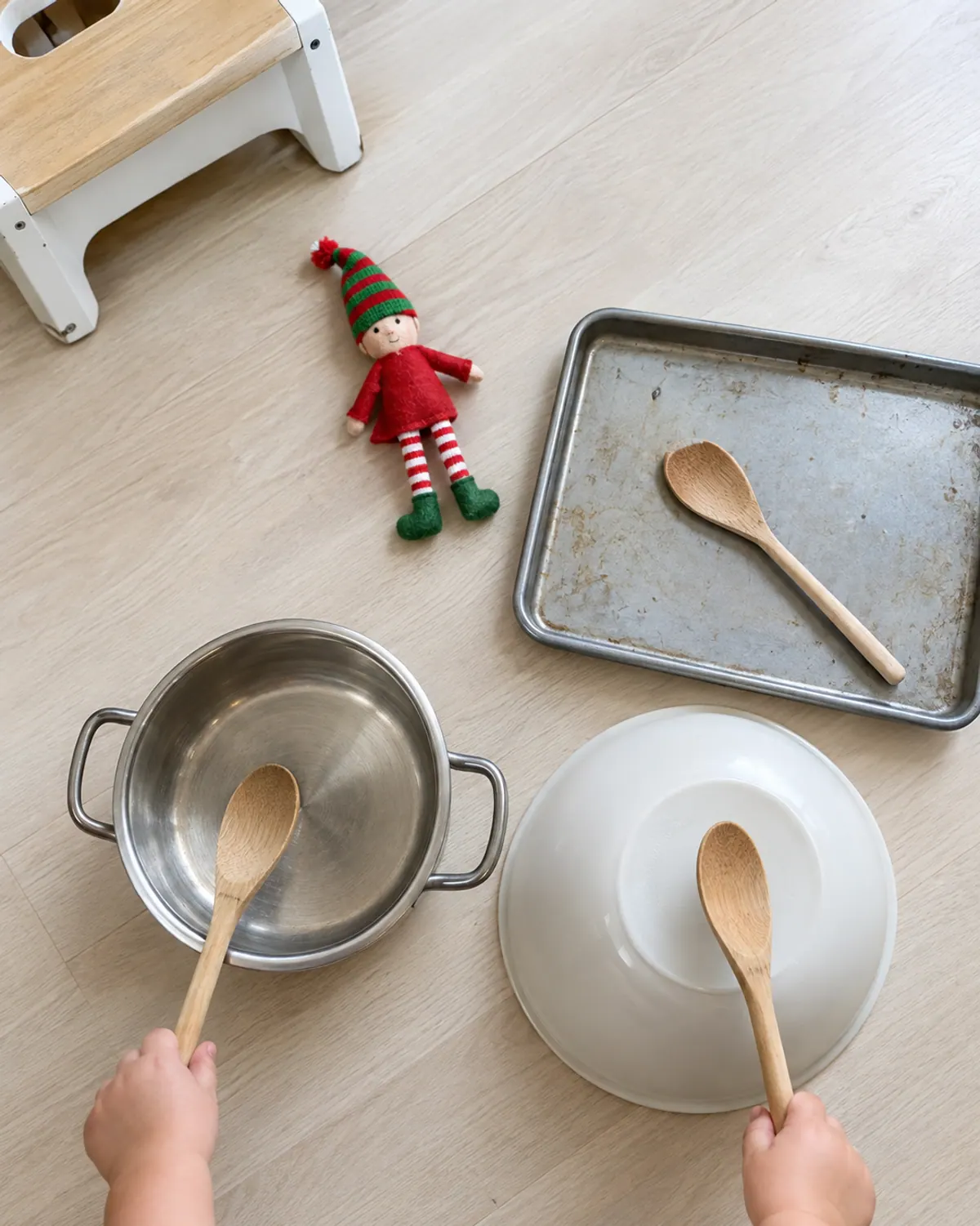 Toddler hand holding a wooden spoon above a pot and baking tray on a kitchen floor, a felt elf doll on a step stool nearby