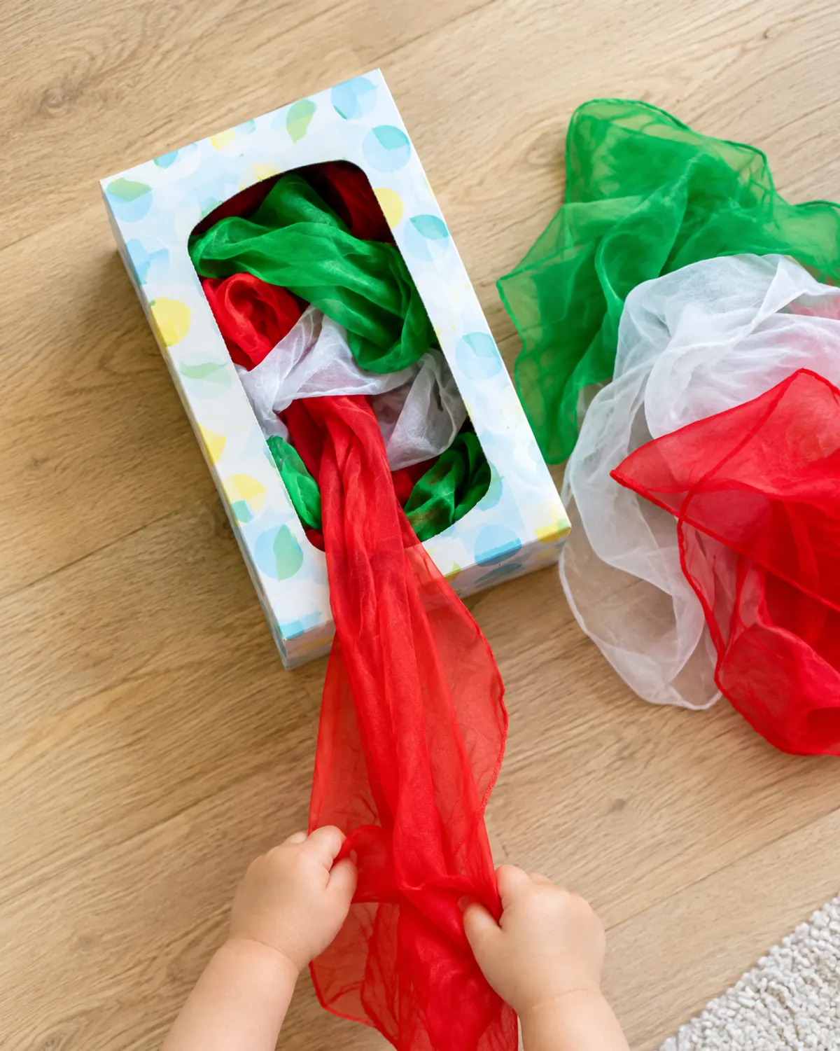 Toddler hands pulling red and green play scarves out of a tissue box, a touchable elf on the shelf idea for toddlers