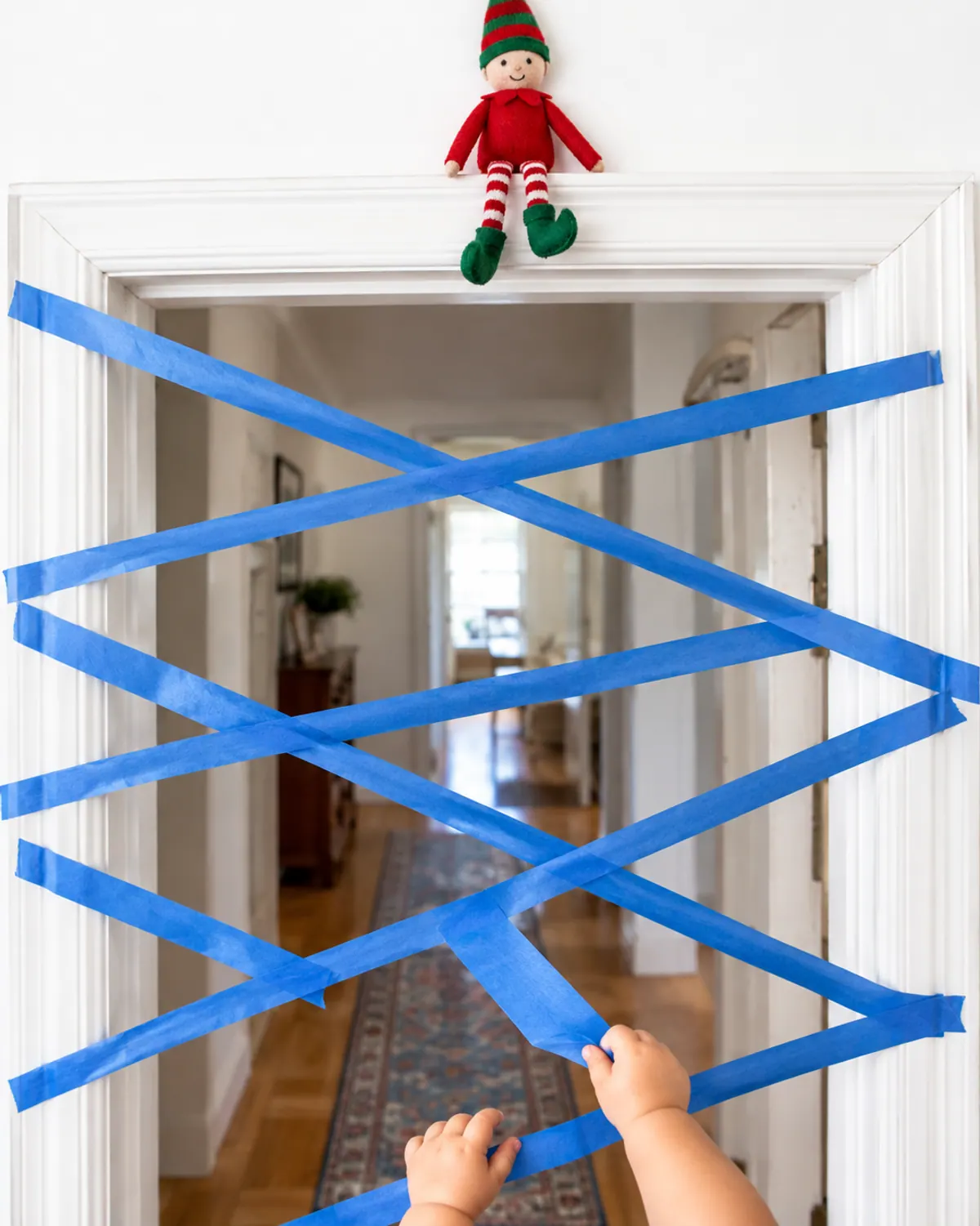 Blue painter's tape web across a bedroom doorway with toddler hands pulling a strip and an elf doll sitting on the door frame above