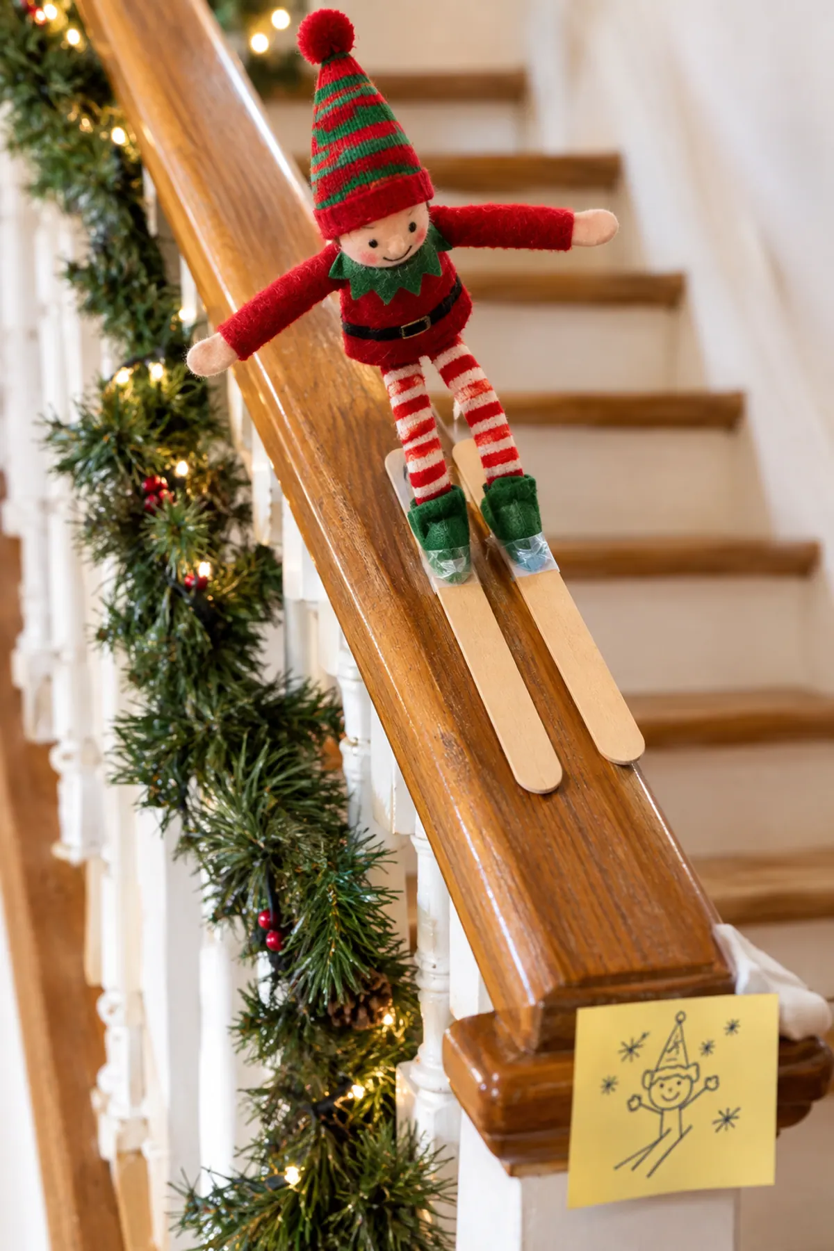 Funny elf on the shelf ideas: a red felt elf doll skis down a wooden stair banister on two craft sticks, a sticky note sign taped at the bottom