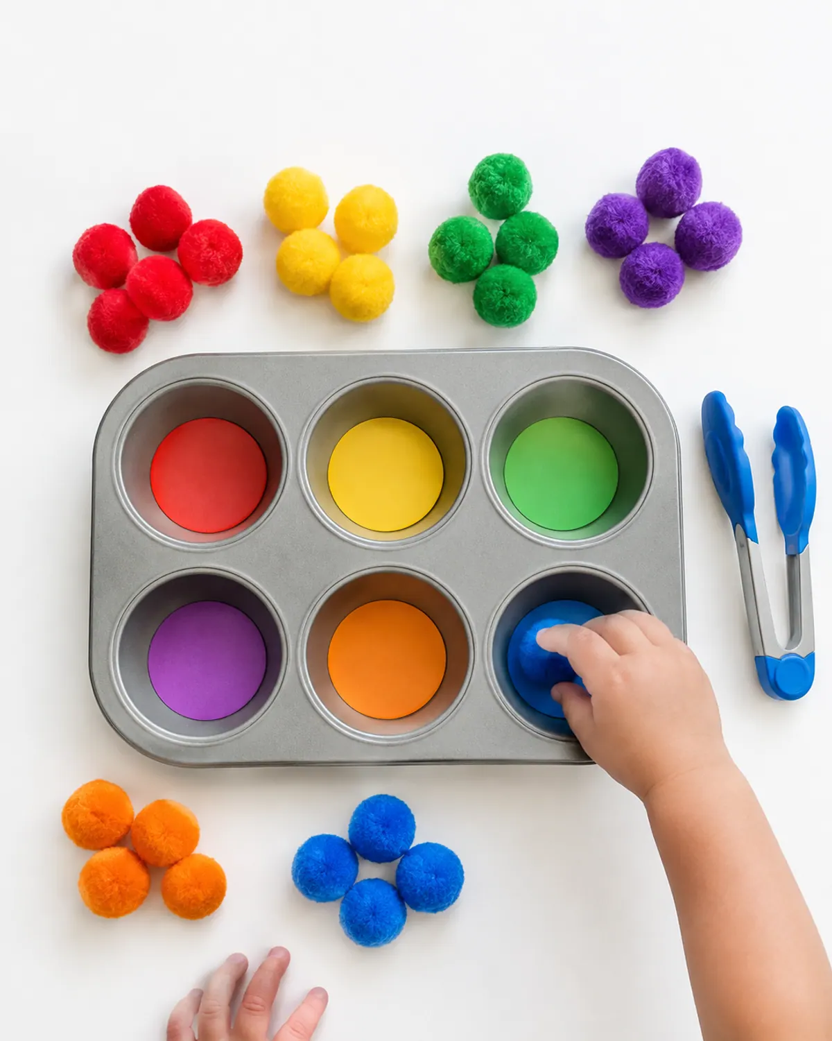 Toddler hand placing a blue jumbo pom pom into a matching blue cup of a muffin tin with chunky tongs nearby