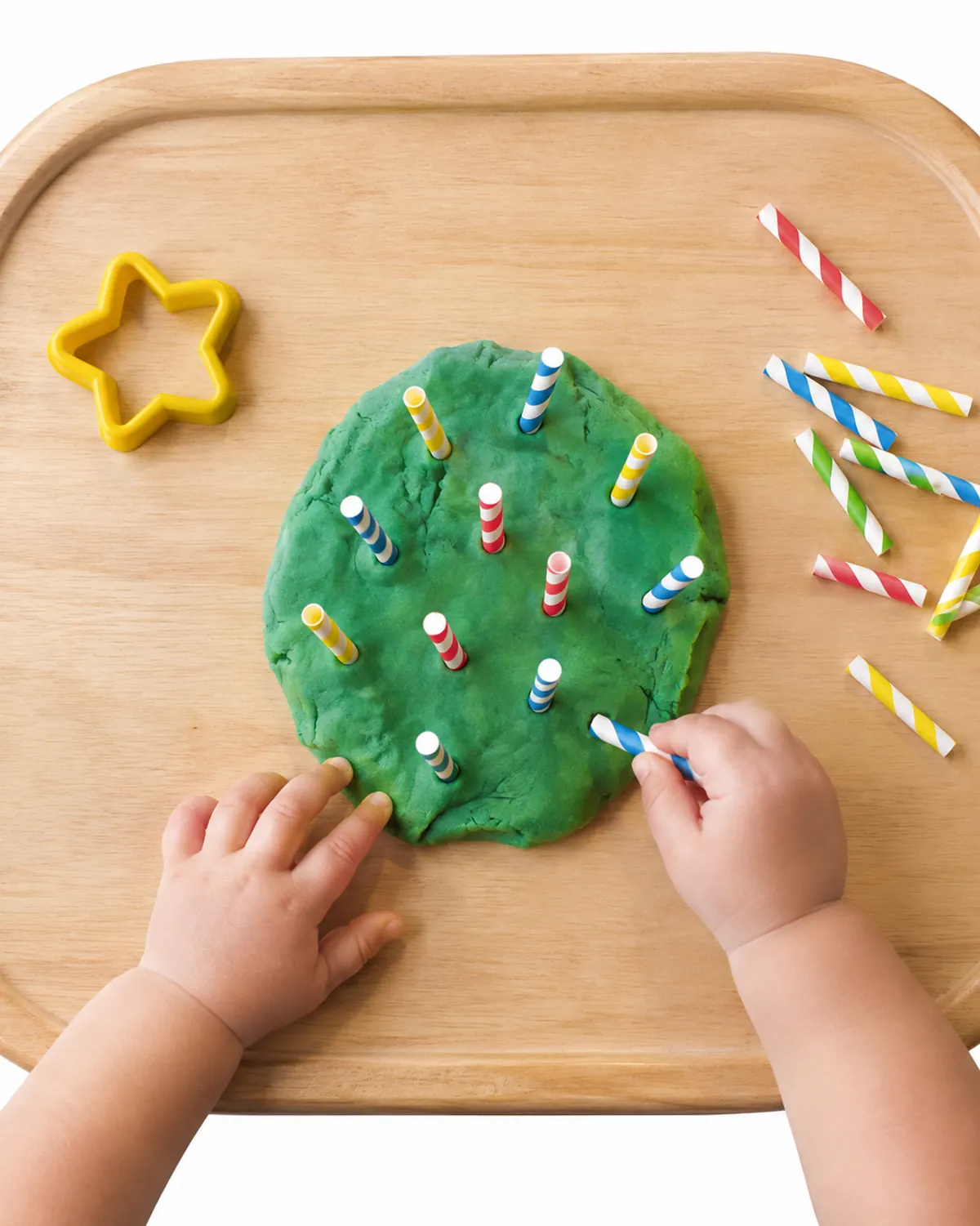 Toddler hand pulling a paper straw out of a flat green playdough cake on a high chair tray