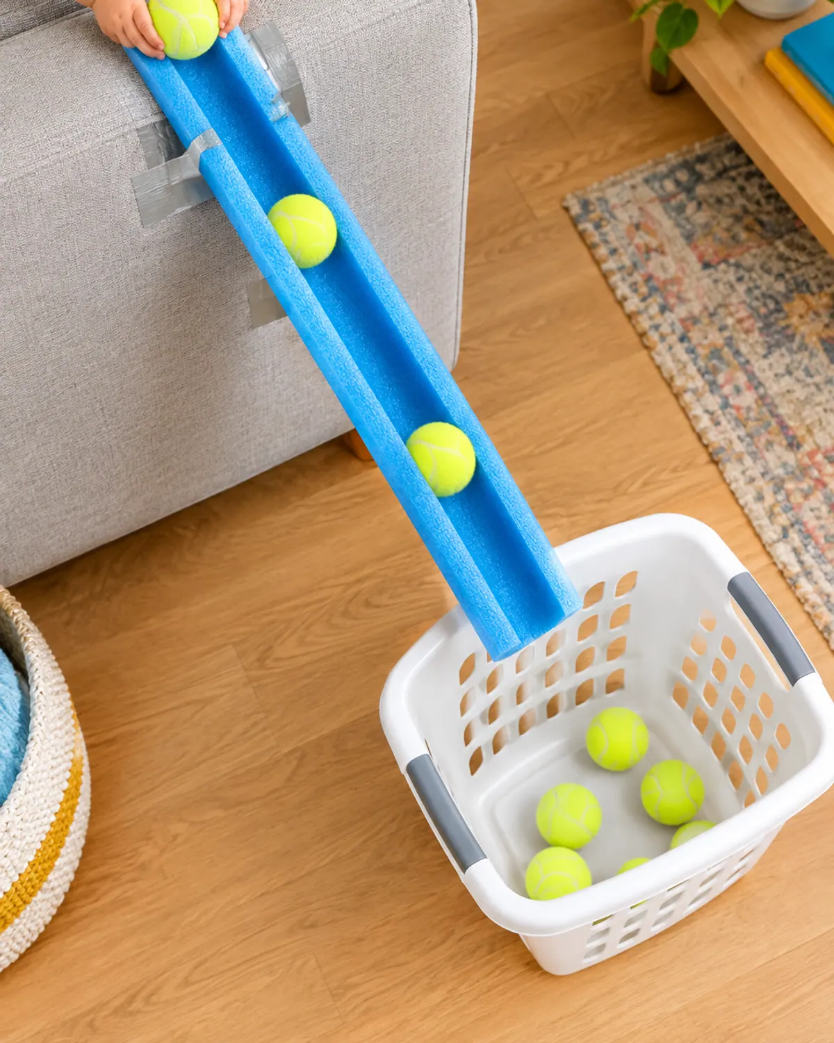 A halved pool noodle taped to a sofa as a ramp with tennis balls rolling into a laundry basket, a toddler hand at the top