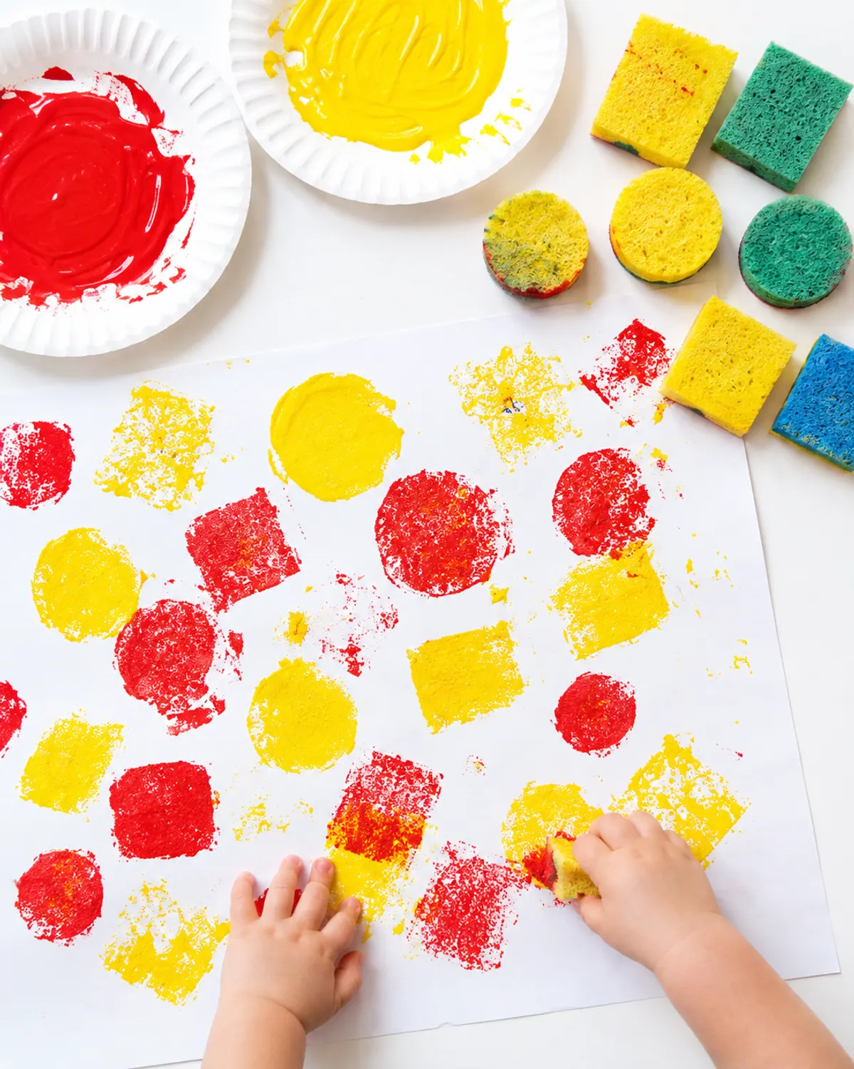 Toddler hand pressing a kitchen sponge in red paint onto paper covered with sponge prints