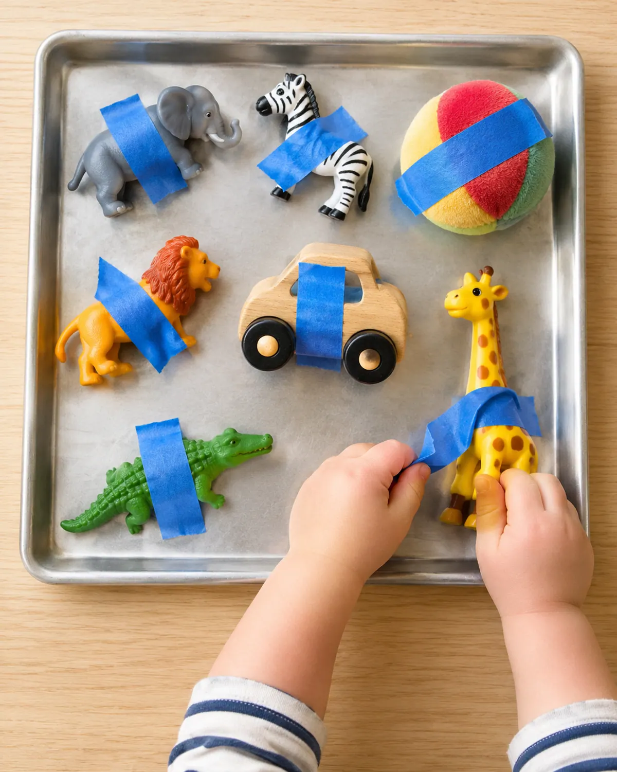 Toddler hand peeling blue painter's tape off a toy giraffe taped to a baking sheet with other toys
