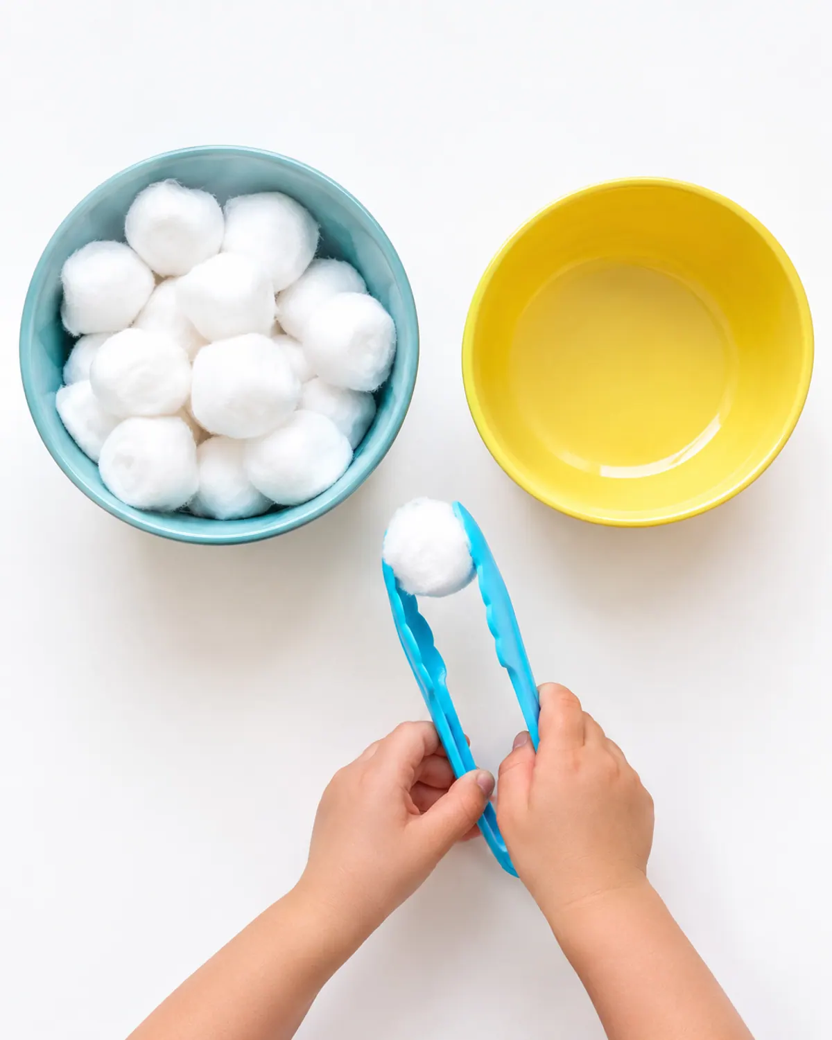 Small hands squeezing kitchen tongs around a cotton ball between two bowls on a wood table