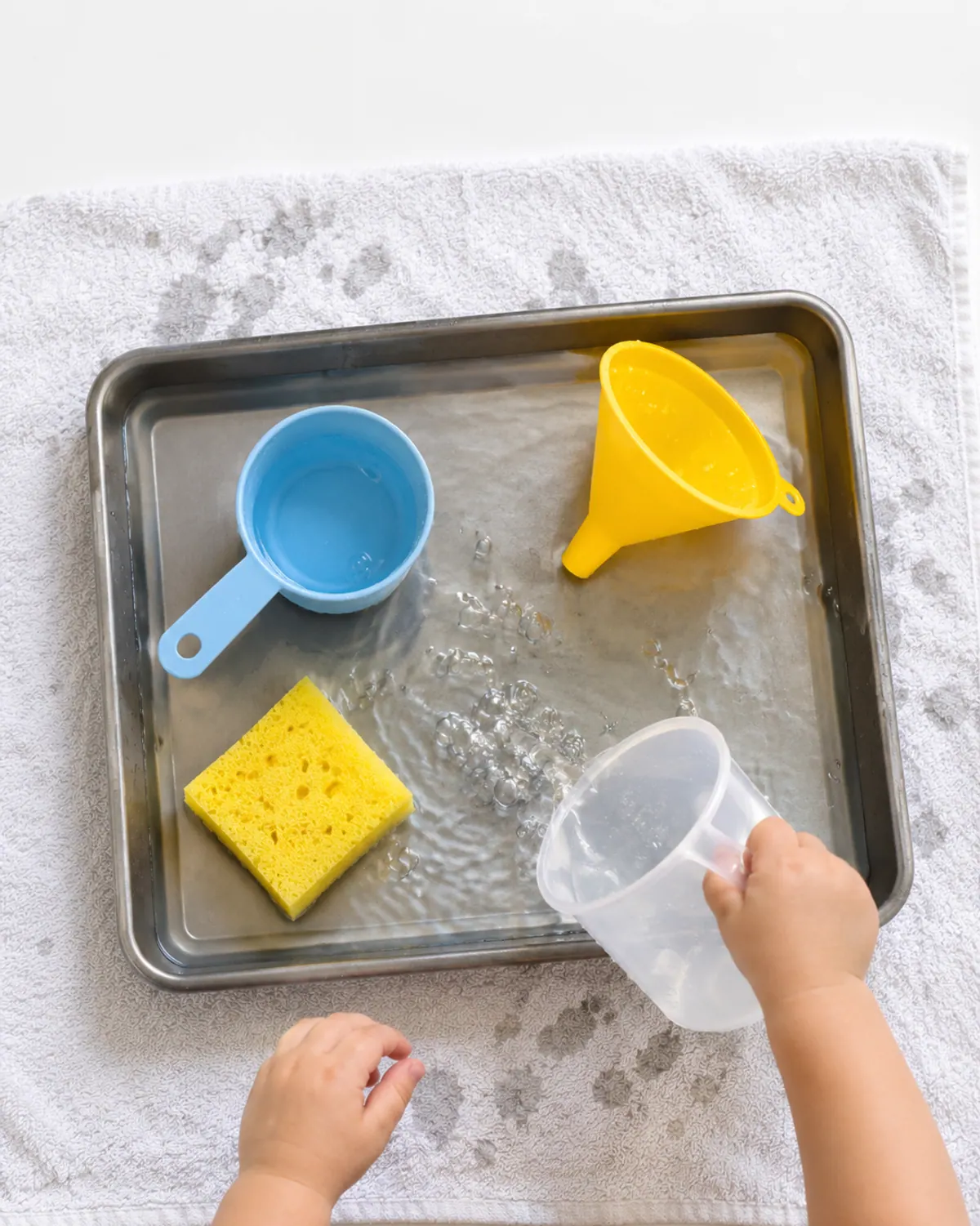 Toddler hands pouring water from a measuring cup into a shallow baking tray on a towel-lined kitchen floor