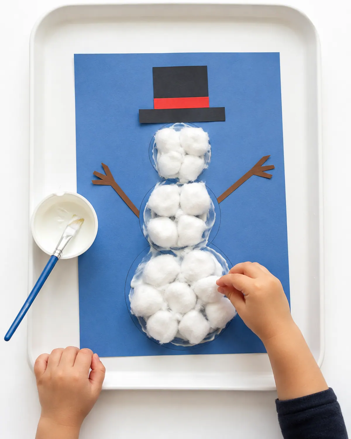 Preschooler hands pressing cotton balls onto glue to make a fluffy snowman on blue paper