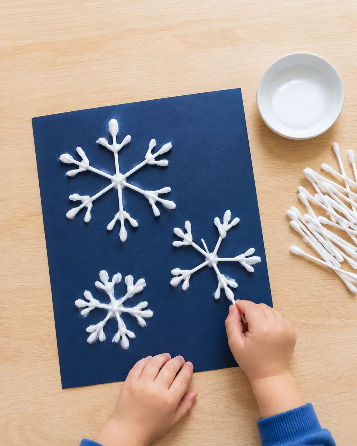 Preschooler hands placing white cotton swabs to form snowflakes on navy blue paper