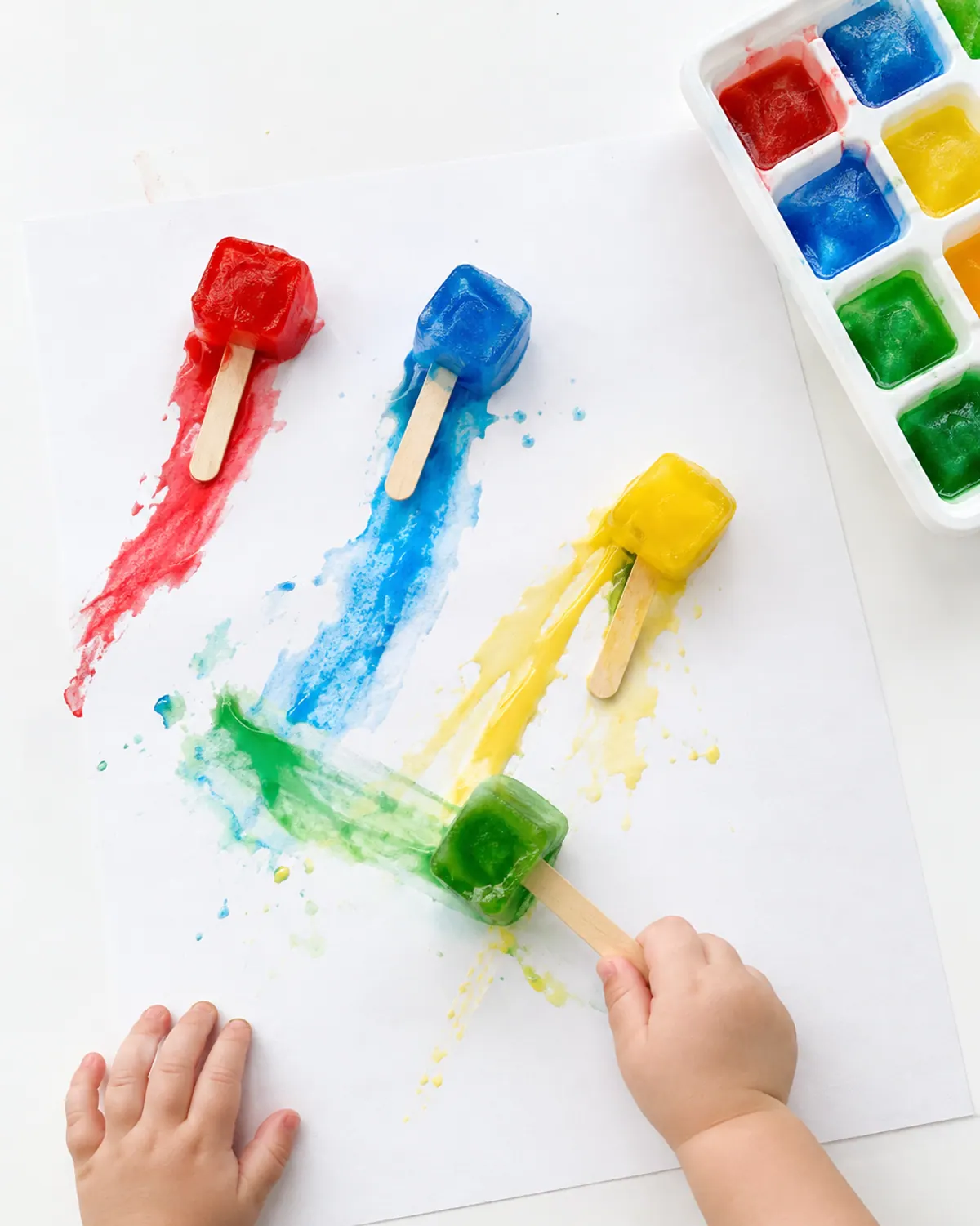 Toddler hand sliding a frozen paint cube on a craft stick across white paper leaving bright streaks