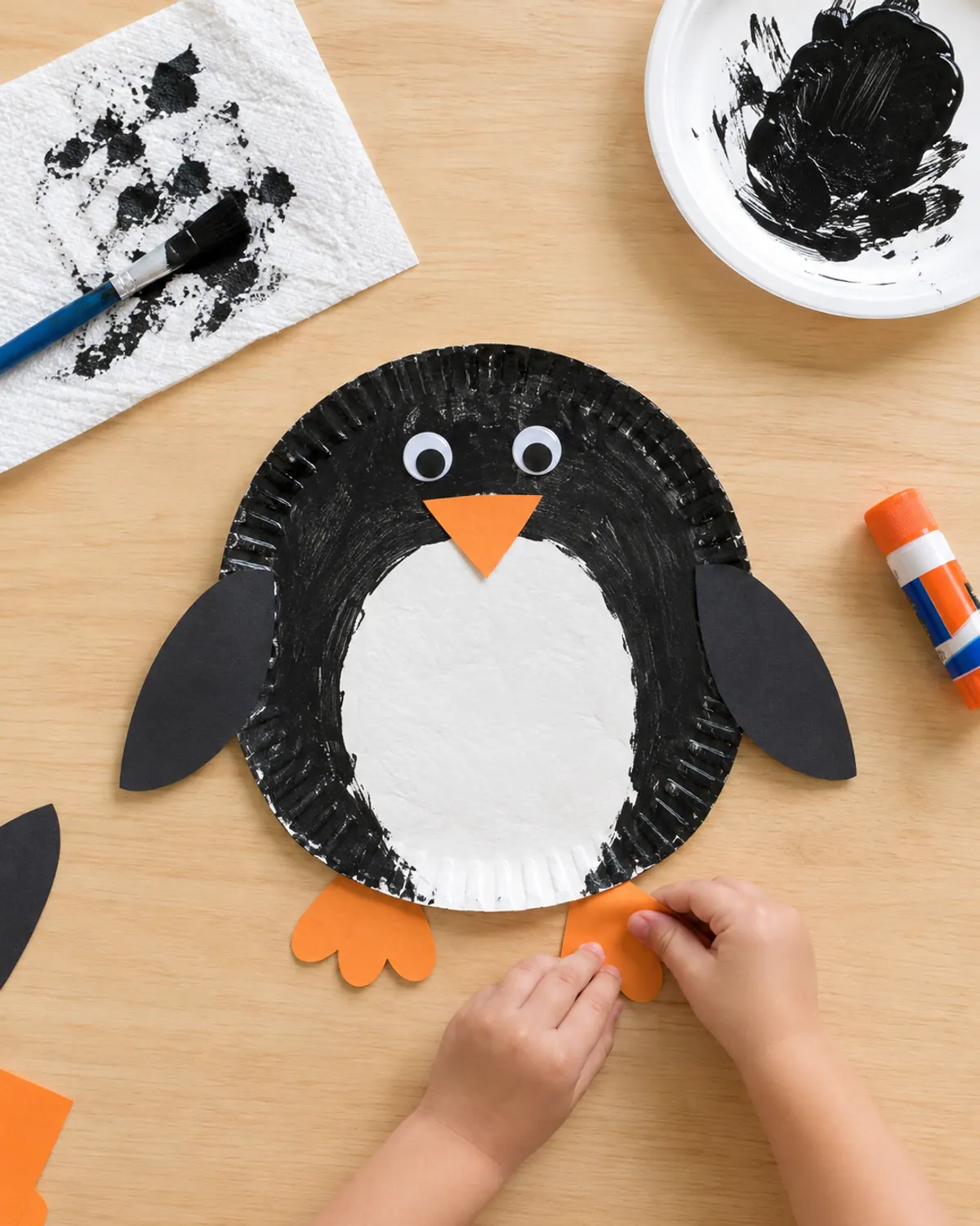 Preschooler hands gluing orange feet onto a paper plate penguin with black painted edges