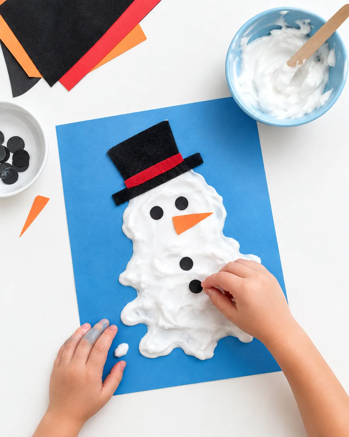 A melted snowman made from white puffy paint on blue cardstock with a felt hat and orange paper nose