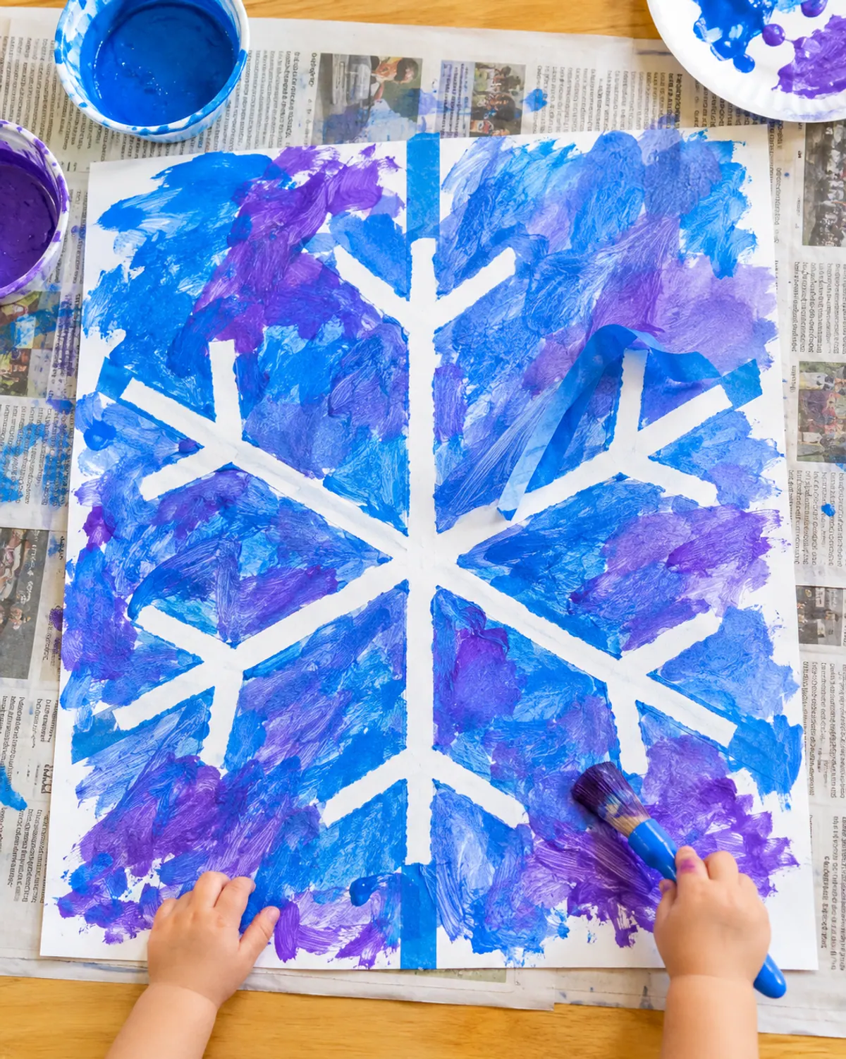 Toddler hand holding a chunky paintbrush next to a tape resist snowflake painting in blue and purple, tape being peeled