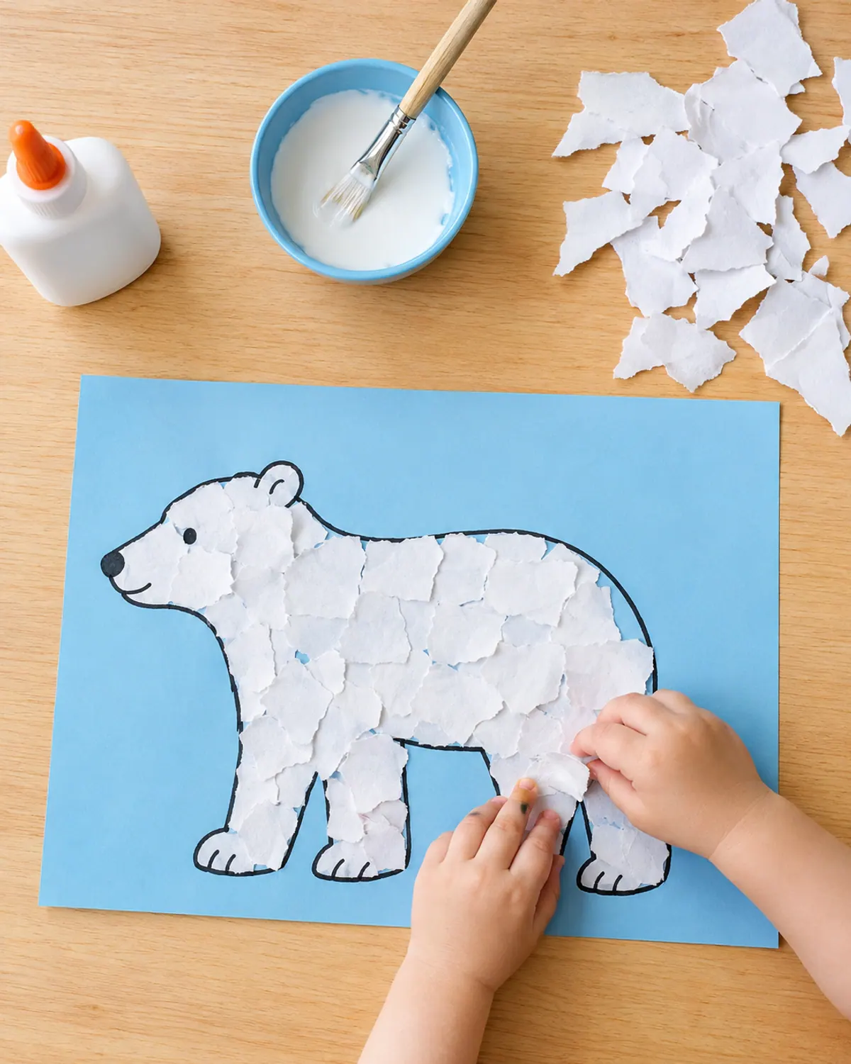 Toddler hands pressing torn white paper pieces onto a polar bear outline on blue paper